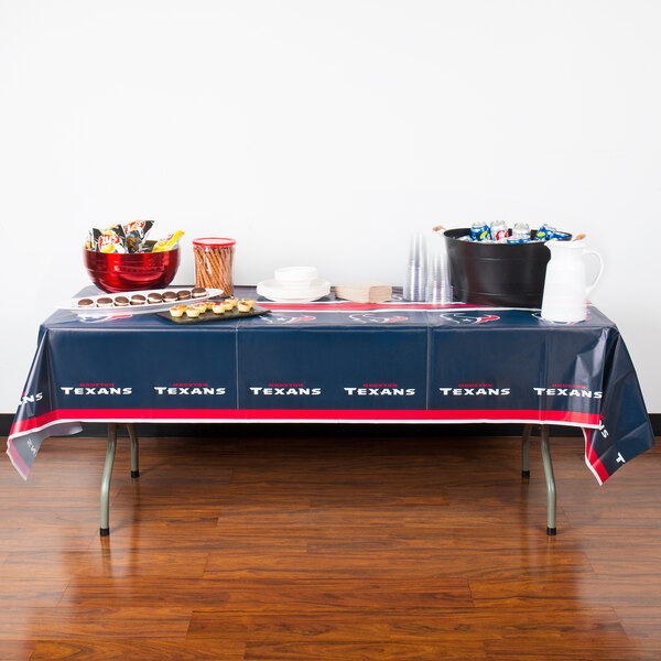 A table with a Houston Texans plastic table cover on it with food and drinks.