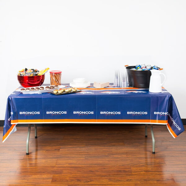 A table with a blue and orange Creative Converting Denver Broncos table cover with food and drinks on it.