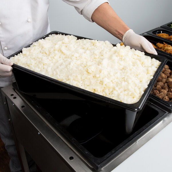 A person in a chef's uniform holding a Cambro black plastic food pan filled with rice.