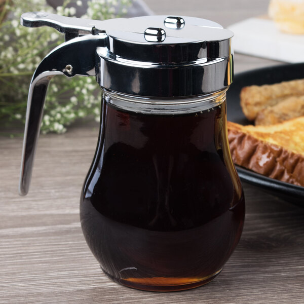 A Tablecraft glass syrup dispenser with a chrome-plated top on a table with a plate of toast.