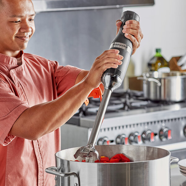 A man using a KitchenAid hand blender to mix red peppers in a pot.