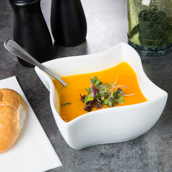 A white American Metalcraft Prestige wave porcelain bowl with yellow soup and a spoon.
