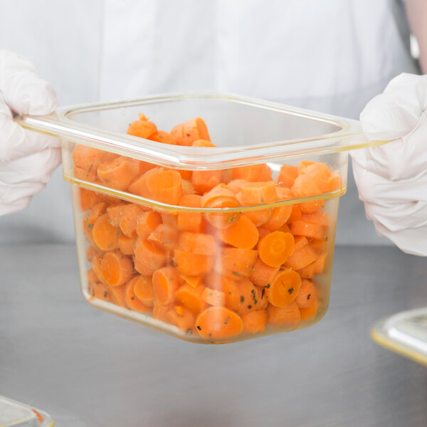 A person in gloves holding a Cambro amber plastic food pan filled with carrots.