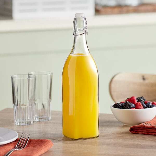 A glass bottle of orange juice with a wire bail swing top lid on a table next to a bowl of fruit.