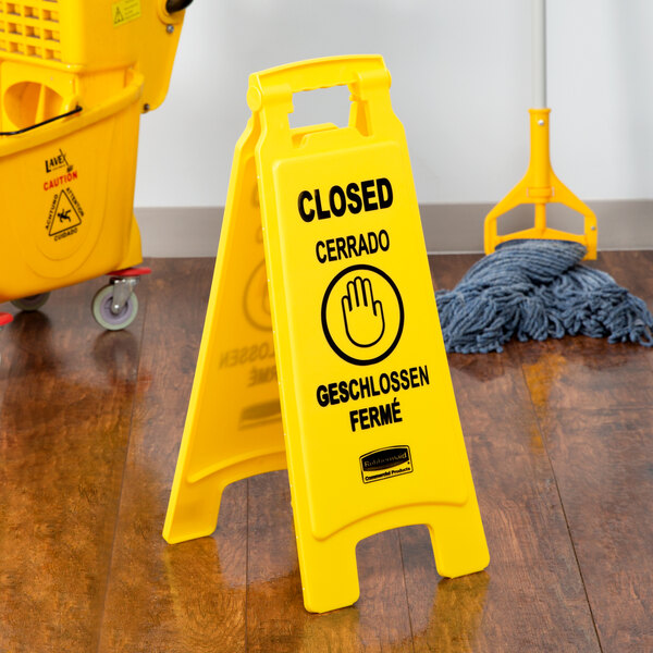 A close-up of a yellow Rubbermaid wet floor sign with a handle and broom and mop icons.