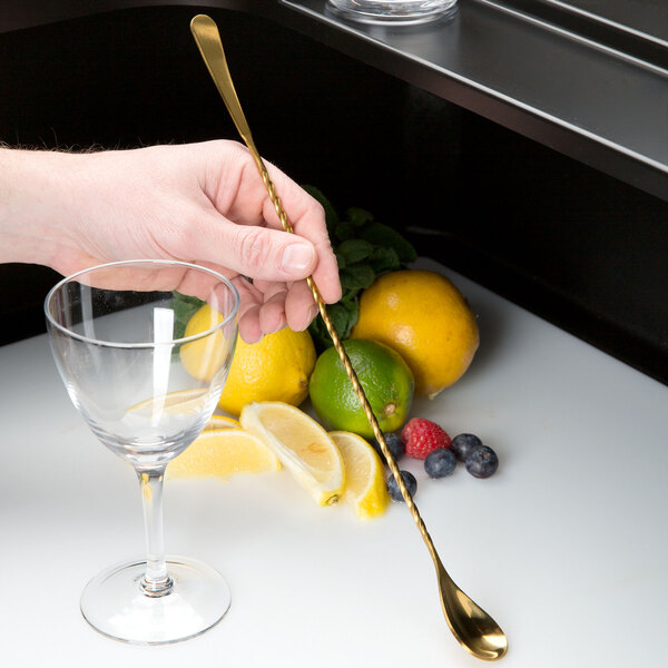 A hand holding a gold Barfly Japanese style bar spoon next to a glass with a lemon slice.