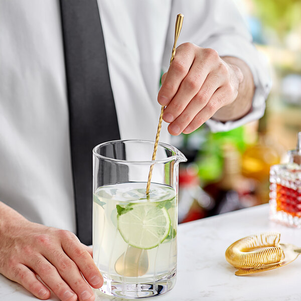 A hand using a Barfly gold-plated classic bar spoon to mix a drink in a glass of water with a lime slice.