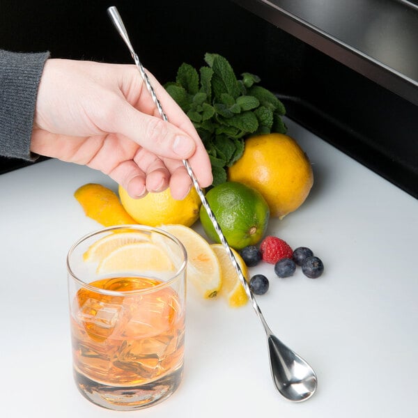 A hand using a Barfly stainless steel classic bar spoon to stir a drink in a glass.