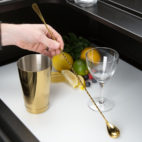 A hand using a Barfly gold plated Japanese style bar spoon to stir a cocktail in a glass with fruit.