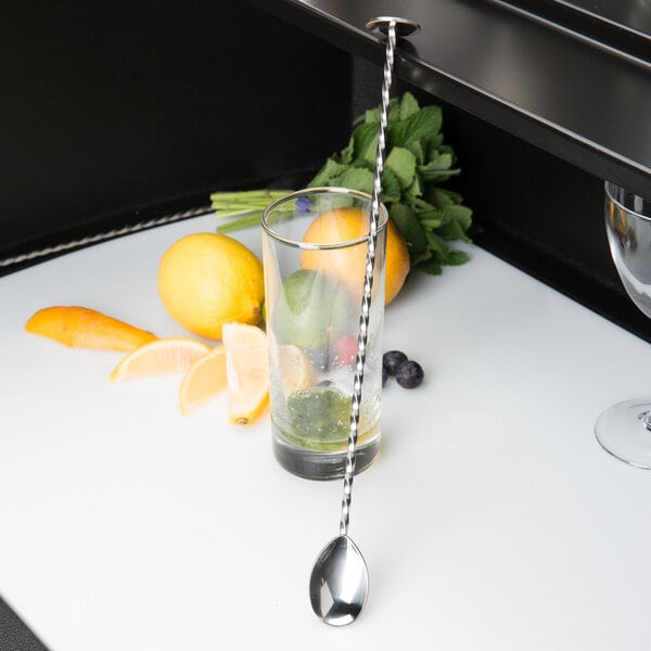 A Barfly stainless steel bar spoon in a glass with fruit on a counter.