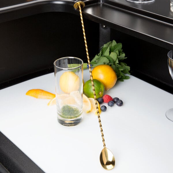 A gold Barfly bar spoon with a muddler end stirring a glass of fruit juice.