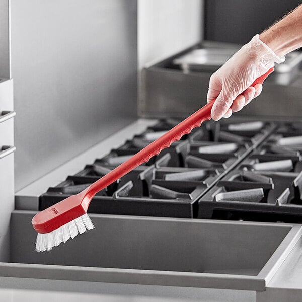 A person in gloves holding a Carlisle red utility brush over a black tray.