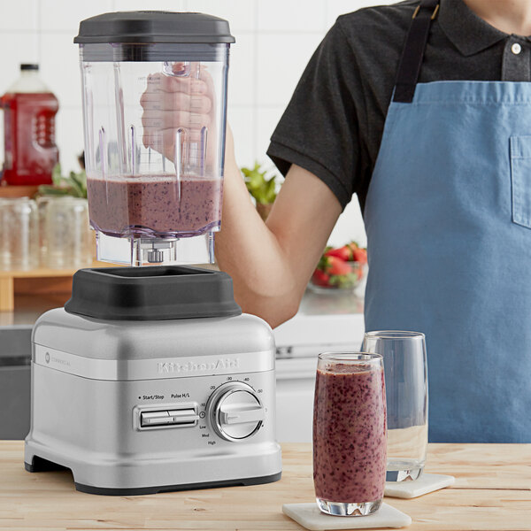 A person in a blue apron using a KitchenAid Contour Silver commercial food blender on a wooden surface to make a smoothie.