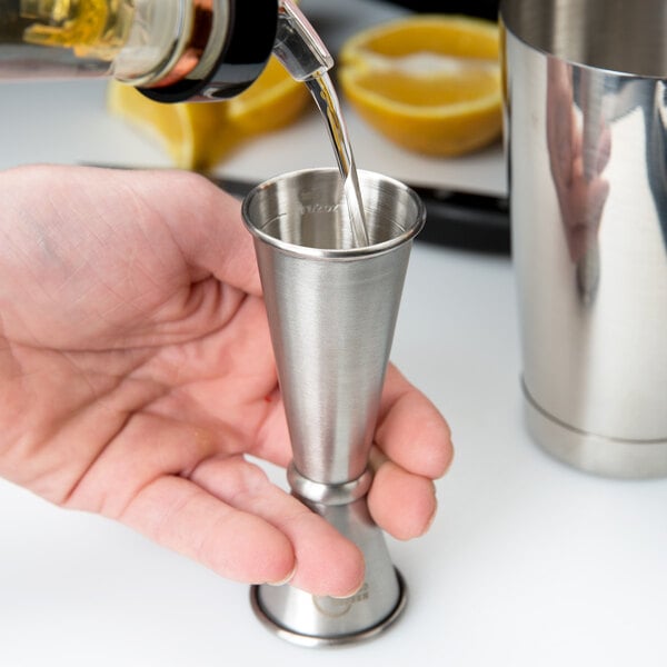 A hand using a Barfly stainless steel jigger to pour liquid into a small metal cup.