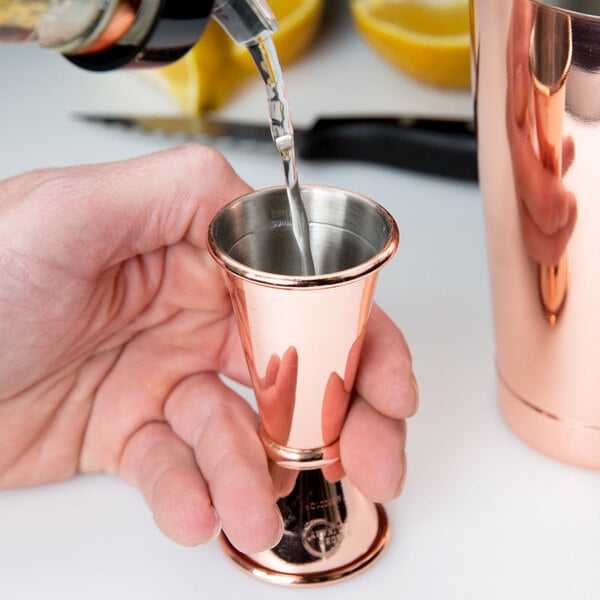 A person using a Barfly copper-plated Japanese style jigger to pour liquid into a copper cup.