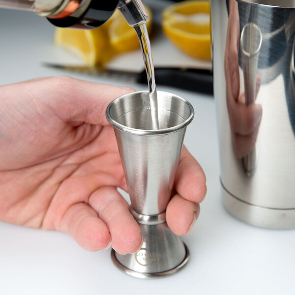 A hand using a Barfly stainless steel Japanese style jigger to pour liquid into a glass.