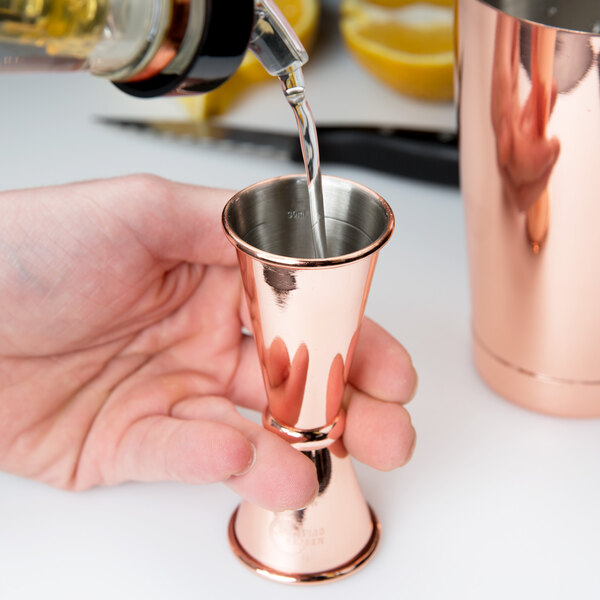 A hand using a Barfly copper Japanese style jigger to pour a shot of liquid into a copper cup.
