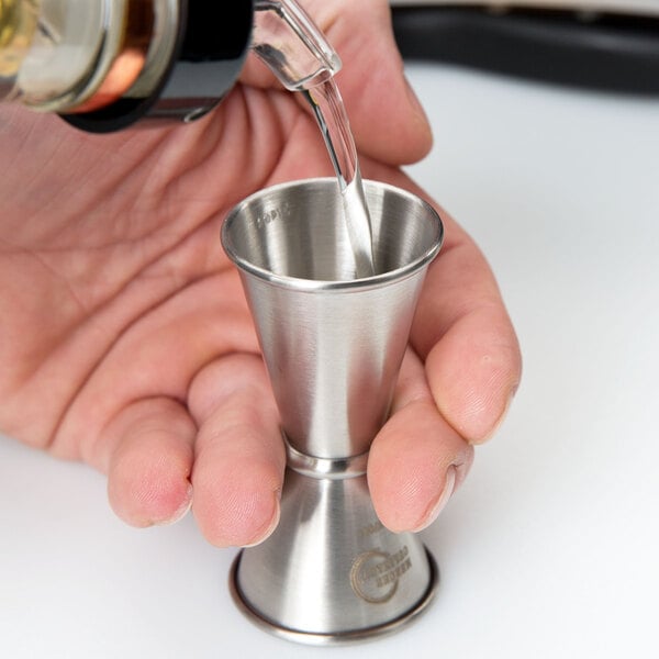 A person using a Barfly stainless steel Japanese style jigger to pour liquid into a small metal cup.