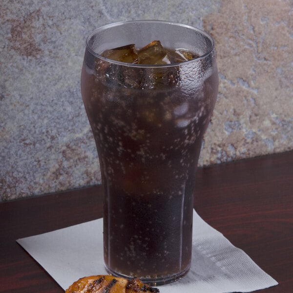 A GET clear plastic soda glass with ice and soda on a table with a napkin and a slice of pizza.