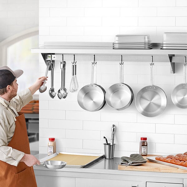 A man in an apron holding a pan and a pan with a pan from the Regency Stainless Steel Wall Mounted Pot Rack.