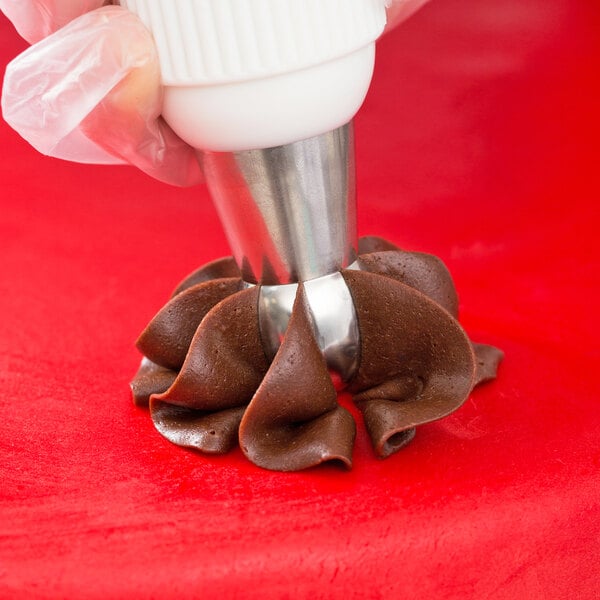 A hand using an Ateco Russian ball tip piping tip to pipe chocolate over cookies on a red surface.