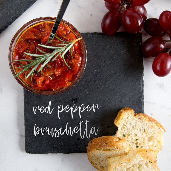 A piece of bread with herbs on a black square plate with a bowl of food and a glass jar of red pepper bruschetta on a counter.