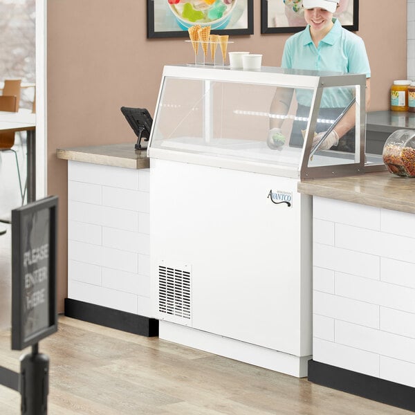 A woman in a white hat working at a refrigerated Avantco ice cream dipping cabinet.