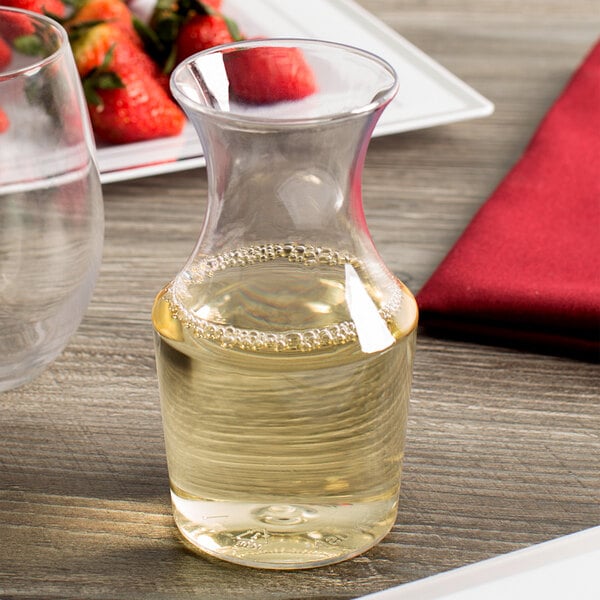A WNA Comet clear plastic wine carafe filled with a clear liquid on a table next to a plate of strawberries.