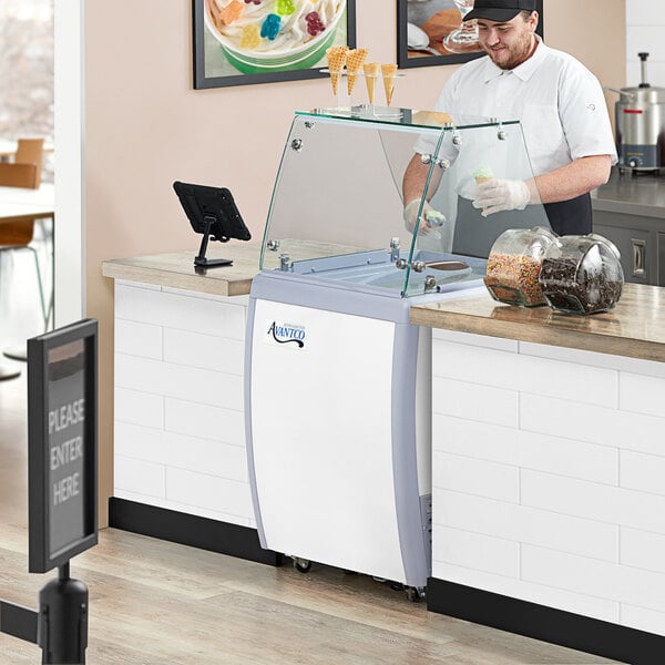 A man in a black hat at an Avantco ice cream dipping cabinet with a glass cover of ice cream.