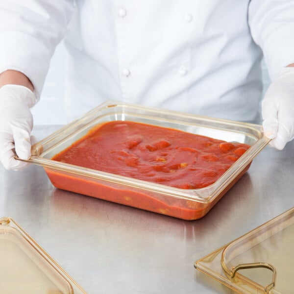 A person in a white coat holding a Carlisle amber plastic food pan with red sauce.