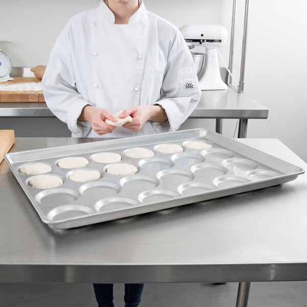 A woman in a chef's uniform holding a Chicago Metallic Aluminized Steel ePan with hamburger buns, muffin tops, and cookies.