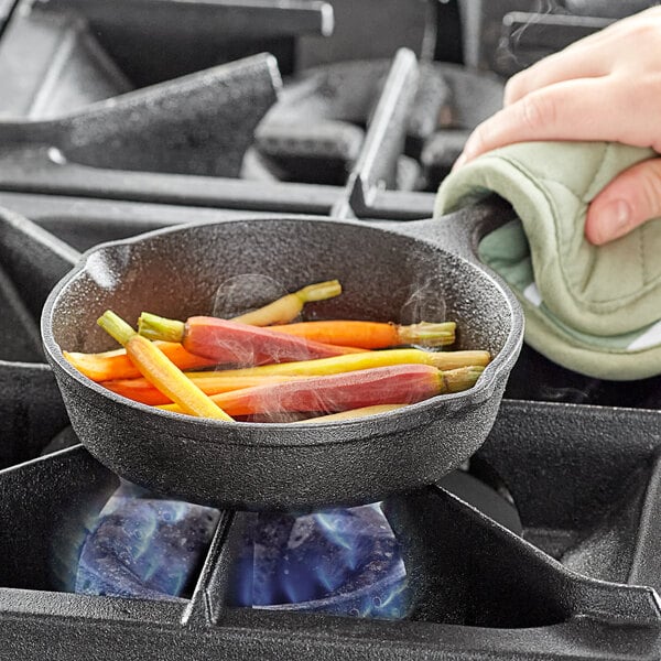 A hand holds a green cloth over a Valor pre-seasoned cast iron skillet with carrots cooking inside.