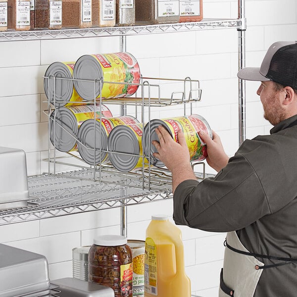 A man in a hat and apron putting cans of tomato sauce into a Regency FIFO wire can rack on a shelf.