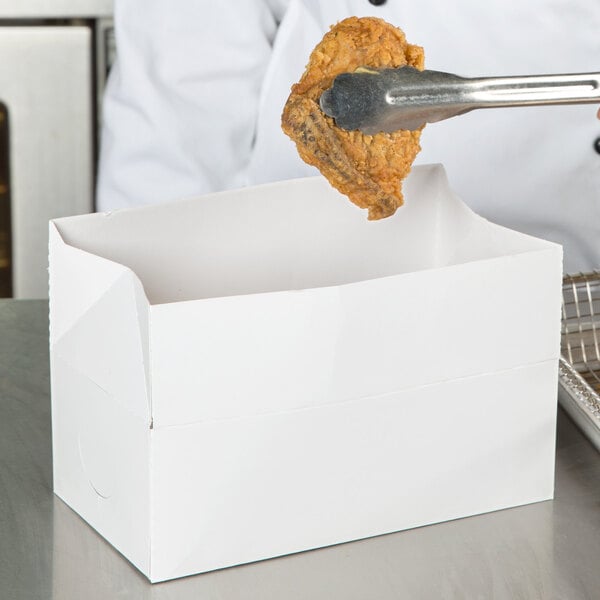 A person holding a piece of fried chicken in a white lunch box.