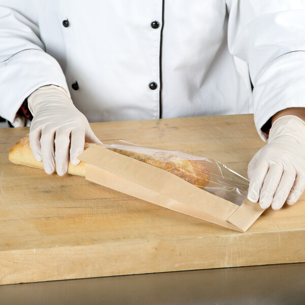 A person in gloves wrapping a baguette in a Bagcraft Packaging brown paper bag with a window.