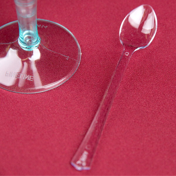 A clear plastic WNA Comet tasting spoon on a red surface next to a clear glass.