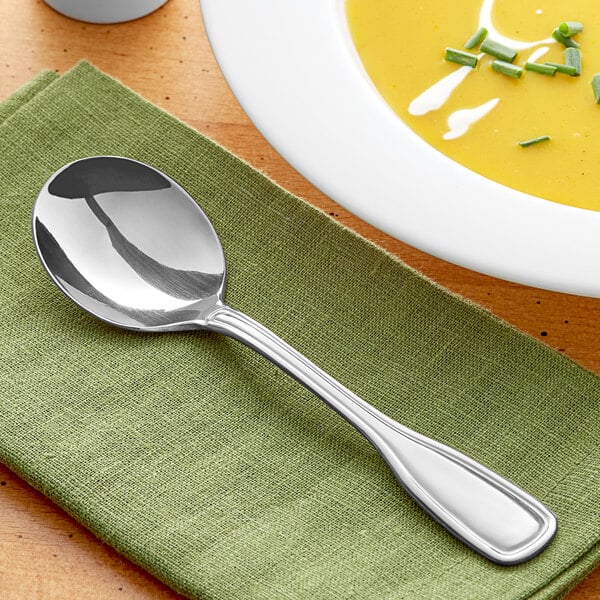 An Acopa Saxton stainless steel bouillon spoon on a green napkin next to a bowl of soup.