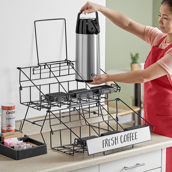 A woman in an apron placing a Choice black wire airpot rack on a counter with a coffee maker inside.