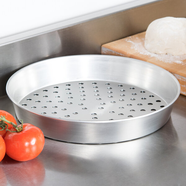 An American Metalcraft tin-plated steel pizza pan with holes next to tomatoes.
