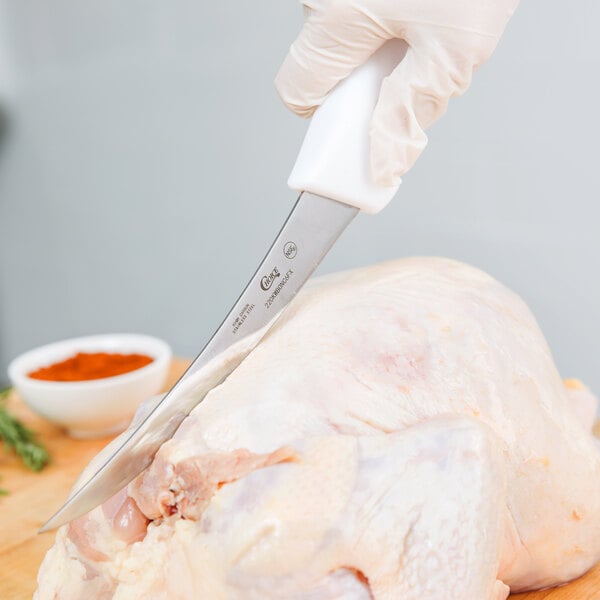 A gloved hand uses a Choice curved boning knife with a white handle to cut a chicken.