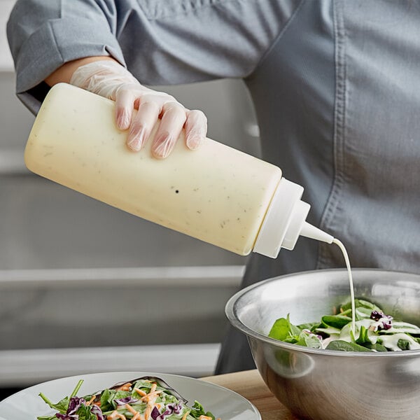A hand using a Choice clear wide mouth squeeze bottle to pour white sauce onto a bowl of salad.