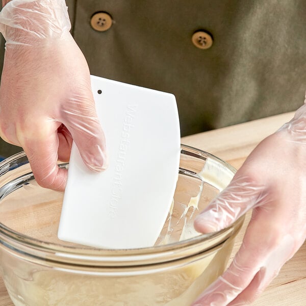 A hand wearing a clear plastic glove uses a WebstaurantStore plastic bowl scraper to mix ingredients in a bowl.