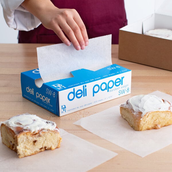A hand using Durable Packaging deli paper to wrap a pastry on a table.