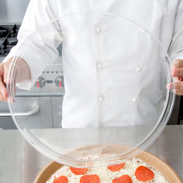 A person wearing a white coat holds a Solut clear plastic lid over a pizza.