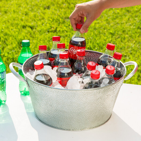 A hand holding a bottle of soda with a red cap placed in a Galvanized Steel Tablecraft Beverage Tub filled with ice.