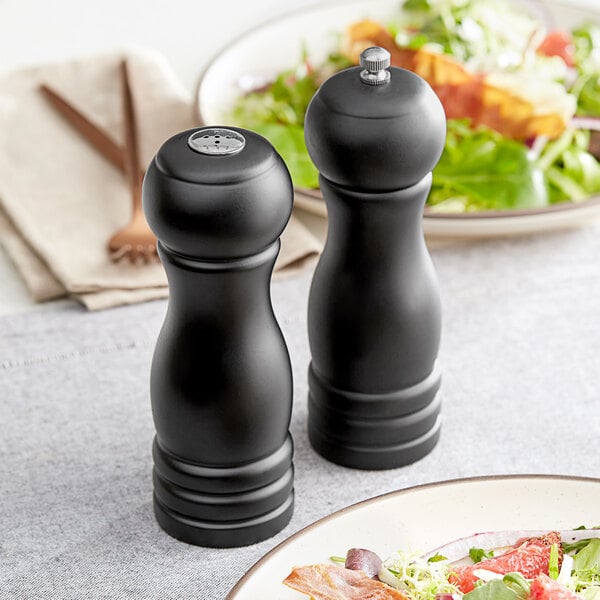 An Acopa matte black wooden salt shaker and pepper mill set on a table with a plate of salad.