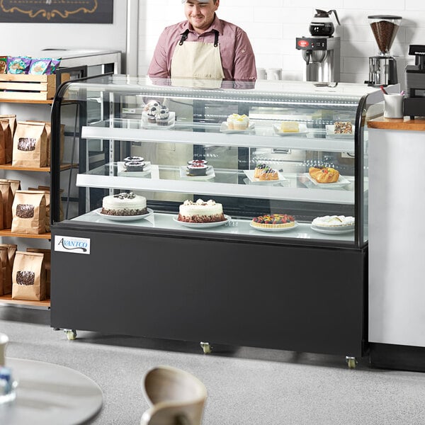 An Avantco curved glass refrigerated bakery display case on a counter with cakes inside, with a man wearing an apron standing behind it.