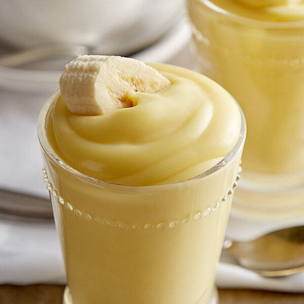 A glass of yellow banana pudding with a white object on top and a banana on the side.