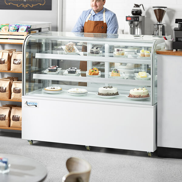 An Avantco refrigerated bakery display case with cakes on a counter with a man wearing an apron standing behind it.