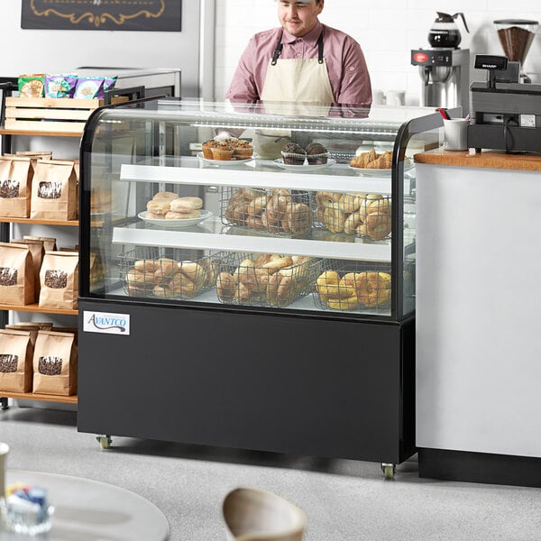 An Avantco curved glass black dry bakery display case on a counter with food and a man wearing an apron standing behind it.
